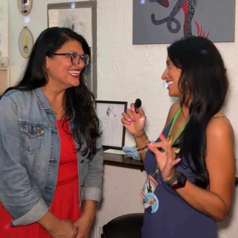 Two women smiling and engaging in conversation at the News Creator Corps event, with artwork and framed pictures on the wall behind them.