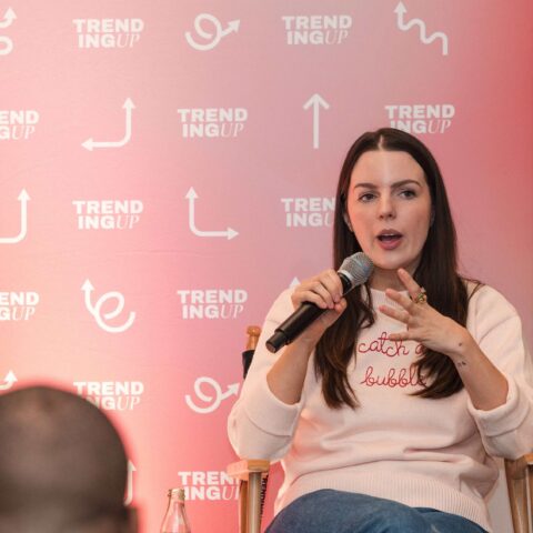 Woman speaking into a microphone at a Trend Up event with a pink backdrop featuring Trend Up logos and arrows.