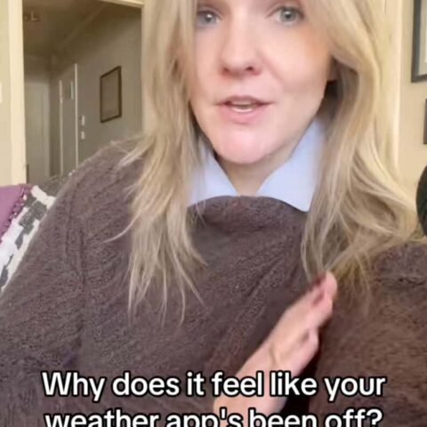 Woman with blonde hair wearing a brown sweater and blue shirt, sitting indoors, asking why her weather app feels off, for News Creator Corps.